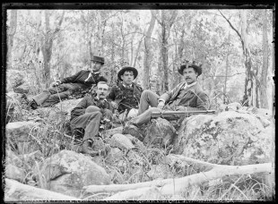 Old time black and white image of four men lying in the forest, one has a pipe, another has leaves around his hat. Museum of Brisbane.