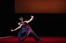 Romeo & Juliet. Two male dancers, onstage against a red backdrop. One is leaning back with their arms open wide, while the other supporting them.