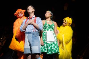 Candide. Four singers lined up. In the middle are a man in blue shirt and shorts with red braces and a woman in a green strappy 1950s type house dress with a little apron. On either side are two women - one all in orange, including her glasses frames and a wig, and the other the same but in yellow.