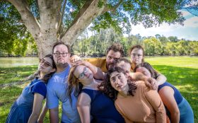 Sprung!! Integrated Dance Theatre Inc, recipient of Create NSW’s Arts and Cultural Funding Program. Photo: Thomas Oliver. Outdoor in a park setting, a group of eight ensemble members from Sprung lean on each other. They appear relaxed.