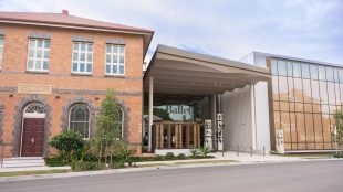 Queensland Ballet. Three buildings next to each other. One a brick two-storey house with five windows and a door, the middle is a recessed glass and concrete ballet centre, and on the right is a large glass facade.