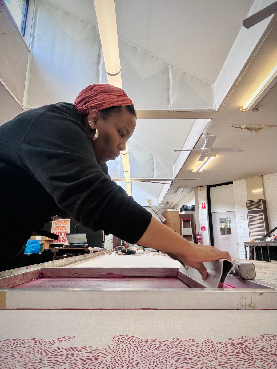 Fashion. A woman of African appearance wearing a pink headscarf works at screen printing on textiles.