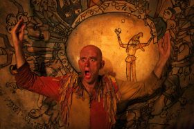 The King's Player. Image is a bald white actor dressed in scruffy Middle Ages clothes with his mouth agape and his arms stretched out in surprise. Behind him is a large yellowish backdrop featuring a circle with a picture of a juggler in it.