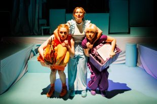 The Roof is Caving In. La Mama Courthouse. Three young women on a stage draped in fabric. In the middle is a woman in white and light blue with her arms around a woman dressed in orange on the left and purple on the right. They are crouching under the middle woman's grasp and hold their belongings in boxes.