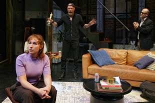 Ulster American. Three actors on a stage set of a loft type apartment. A woman in a short sleeved lilac jumper sits downstage. Behind her centre stage is a bearded man all in black with a baseball cap backwards and an Oscar in his outstretched hand; he is shouting. A third balding man with glasses stands on the right behind a tan leather couch clasping his hands and watching the man in the centre.