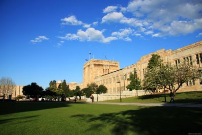 UQ is planning to cut its Museum Studies program. Image is an exterior shot of the sandstone façade of Queensland university, behind some trees, in long shot against a blue sky with scudding clouds.
