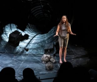 World Problems. Image is a woman in grey standing on a stage in front of the mouth of a tunnel, that looks as if it's lined in slate. There are boulders at her feet and we can see the sihouetted heads of the front row of the audience from behind.