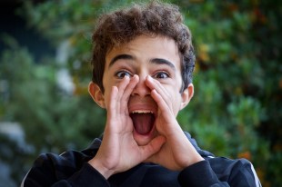 arts news. image is a young boy standing in front of foliage with his hands cupping his mouth to shout.