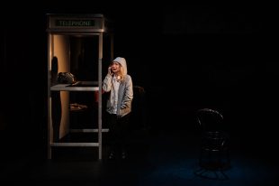 WAY. On a dark stage an older worried looking white woman in a grey hoodie is using a phone in a telephone box. A chair is on the right of the picture.