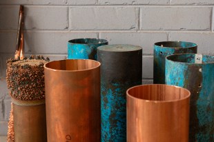 Canberra Museum and Gallery. A group of metal cylinders in shades of bronze and blue patina are grouped against a white brick wall. The one on the far left is bubbly at the top as if it has been underwater or in an acid bath.