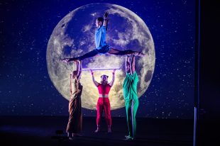 I Wish... In front of a backdrop of a huge moon in the night sky, four circus performers dressed in yellow, blue, red and green perform, with two performers holding aloft a third who is doing the splits, while the fourth performer stands at the back holding up an Astera light tube.