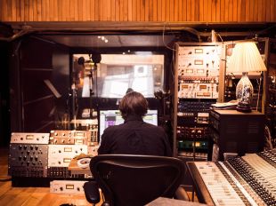 A young man sitting in a music recording studio surrounded by sound equipment.