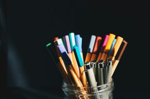 Handwriting. Typing. Image is a glass jar against a black background, filled with a variety of pens and pencils.