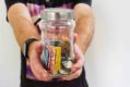 A glass jar containing wads of Australian dollars and coins is held towards the camera by an anonymous figure.