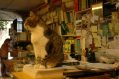 cat in a book. Tabby cat sits in a studio on a pile of papers or manuscripts. It is facing the left and looking off into the distance. The studio is filled with books and detritus and there is a woman in the background doing some sort of admin.