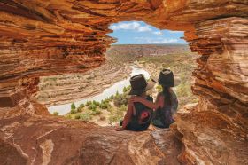 A natural rock formation in the rough shape of a window looks out over a river and surrounding Nanda Country. Two children wearing Akubra hats sit in the window, their backs to the camera.