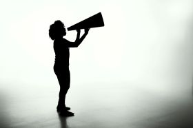 A black and white photograph of a woman with an old-fashioned megaphone silhouetted in profile against a white background.