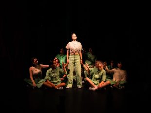 The Word. On a dark stage a group of young people sit in a huddle. They wear shades of green and are turned to a young woman in the middle who is standing. Some of them point at her.