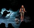 World Problems. Image is a woman in grey standing on a stage in front of the mouth of a tunnel, that looks as if it's lined in slate. There are boulders at her feet and we can see the silhouetted heads of the front row of the audience from behind.