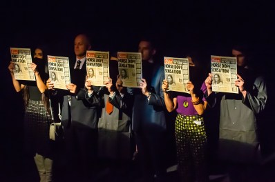 Ink by New Theatre. Six people are lined up on a dark stage holding identical copies of The Sun newspaper in front of themselves, obscuring their faces.