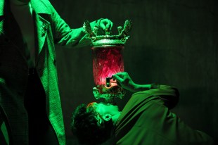 Death in the Pantheon. On a dark stage two actors are bathed in green light. One can only be seen from the neck down and is holding an ornate drinking contraption, with a red glass case, the other is bending below it to drink.