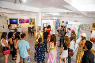 A man in black is in the centre of a room surrounded by people. He's standing by a painting. There are other paintings around the room.