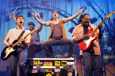 RISING: A group of three First Peoples and a Caucasian man performing in a rock band with an arch of stage lights behind them. Two are playing guitars, the lead singer is standing on a packing case and one is waving drum sticks.