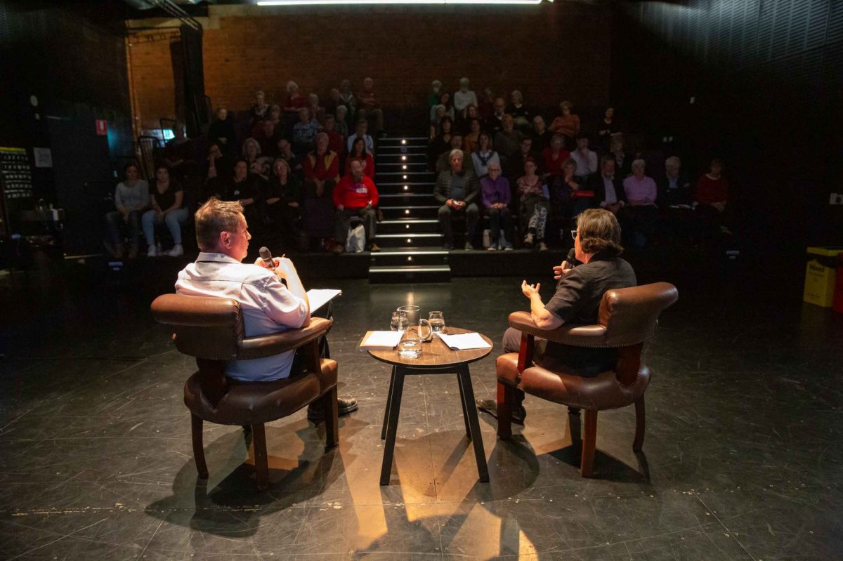 Two people are sitting facing the audience with their backs to the viewer. There is a table between them. They are in mid-conversation.