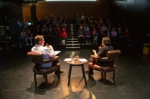 Two people are sitting facing the audience with their backs to the viewer. There is a table between them. They are in mid-conversation.