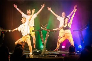 Three men and a woman are standing on treadmills. They have their arms in the air.
