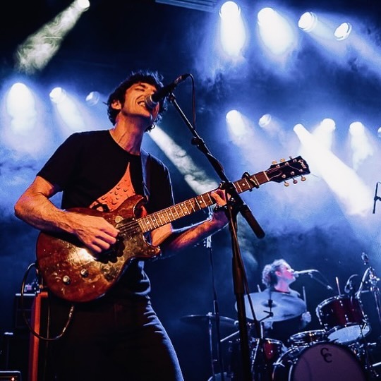 A dynamic photo of a two-piece punk band playing live. A dark haired man in a black t-shirt plays the guitar as he sings into a microphone; behind him a woman plays drums.