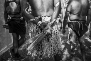 A black and white photograph of three members of the Deaf Indigenous Dance Group, all of whom stand with their backs to the camera. The photo is focused on a central figure who wears a grass skirt and holds clapping stocks behind his back. A figure in shorts stands to his left, and another figure in a loincloth stands to his right.