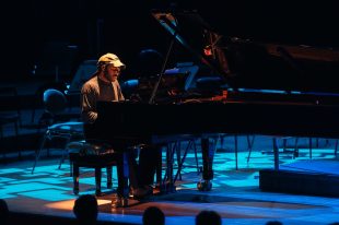 Devonté Hynes. An African American man with a black short beard, brown cap and brown jumper sits at a grand piano playing intently.