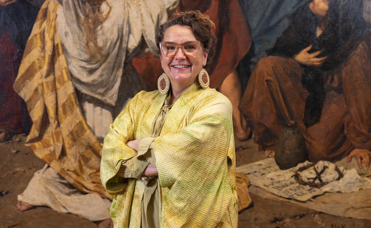 Woman with yellow shirt, glasses, and big earrings, smiling, in front of Aboriginal painting. Lisa Slade.
