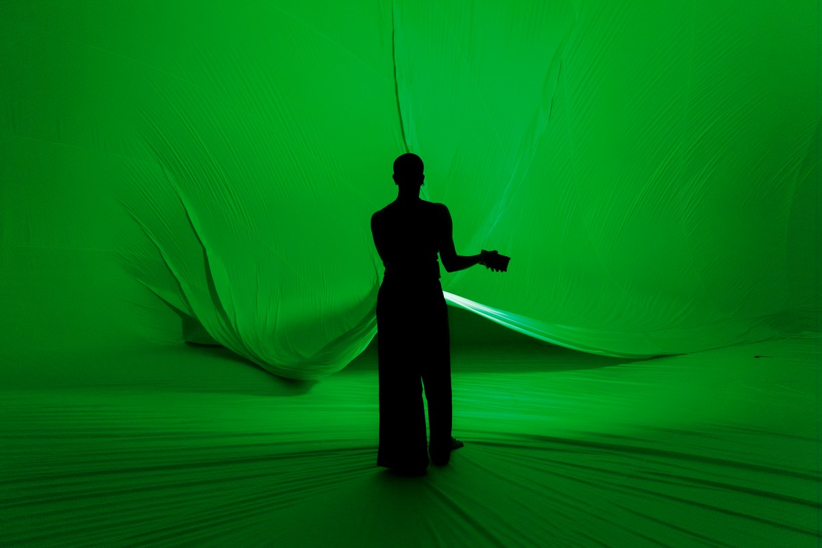 RISING: A silhouette of a shaven headed dancer holding out a green light and standing with their back to the camera in front of a large inflatable.