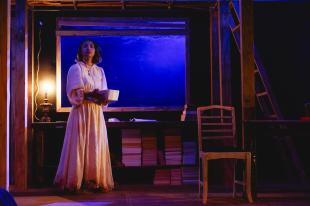 Ghosts. A young woman in a white dress stands on stage in a dark and gloomy house set with a blue/purple light behind where through the window. There is a lonely chair and piles of books near her.