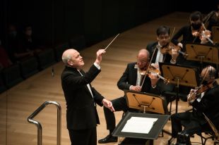 Standing at a podium, his eyes closed in concentration, a bald-headed white man wearing a black suit and white shirt conducts an orchestra.