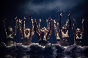 Ten figures are sitting cross-legged in formation with their hands raised above their heads. They are wearing light, floaty trousers. The men are bare-chested. The women are wearing brown-coloured bras.