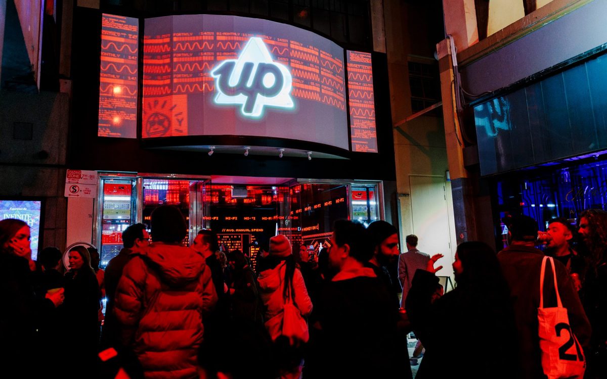 Night Trade at RISING 2024. Photo: Gianna Rizzo. People gathering in an alleyway where a bit screen is showing a white triangle with the word ‘up’. The image is saturated by red light.