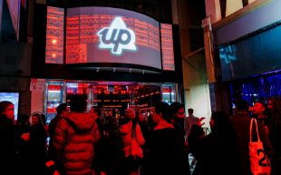 Night Trade at RISING 2024. Photo: Gianna Rizzo. People gathering in an alleyway where a bit screen is showing a white triangle with the word ‘up’. The image is saturated by red light.