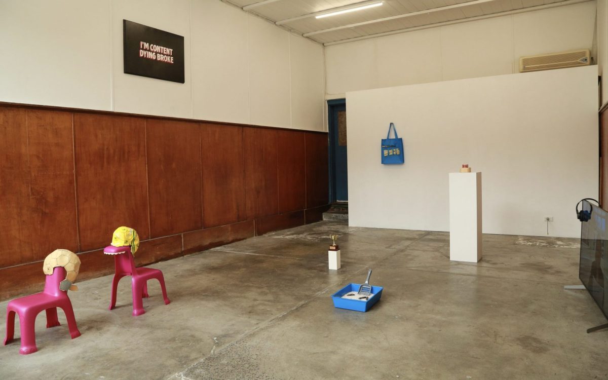 ‘Joke Taxonomy’, installation view at 138 Gallery. Photo: Supplied. A brown wooden panelled gallery space with readymade objects including a pair of pink Dino chairs, a litter box, and a small trophy.