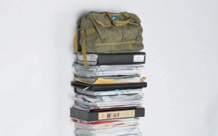 Napier Waller Art Prize-winner 2024 Kat Rae, ‘Deathmin’ (detail), 2023, stacked paper, vinyl, plastic, leather, metal. Image: Supplied. Folders of paperwork stacked on top of each other against the wall, with a green army bag placed on top.