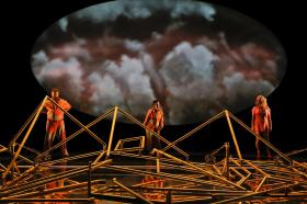 Three actors in orange hi-vis clothes with a huge oval backdrop showing clouds, stand behind a mass of complicated poles.