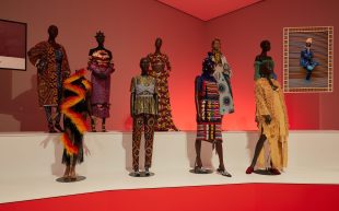 Installation view of 'Africa Fashion' on display from 31 May to 6 October 2024 at NGV International, Melbourne. Photo: Lillie Thompson. Nine mannequins standing on a tiered display with red lights. Each of them are wearing glittery and glamorous garments by African fashion designers.