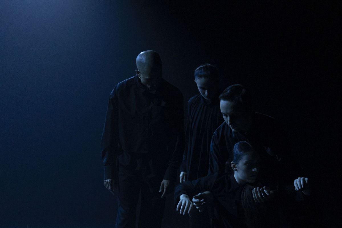 A darkened stage with four performers dressed in black in a simple formation, some are on the floor, others are standing.