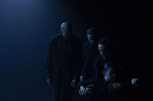 A darkened stage with four performers dressed in black in a simple formation, some are on the floor, others are standing.