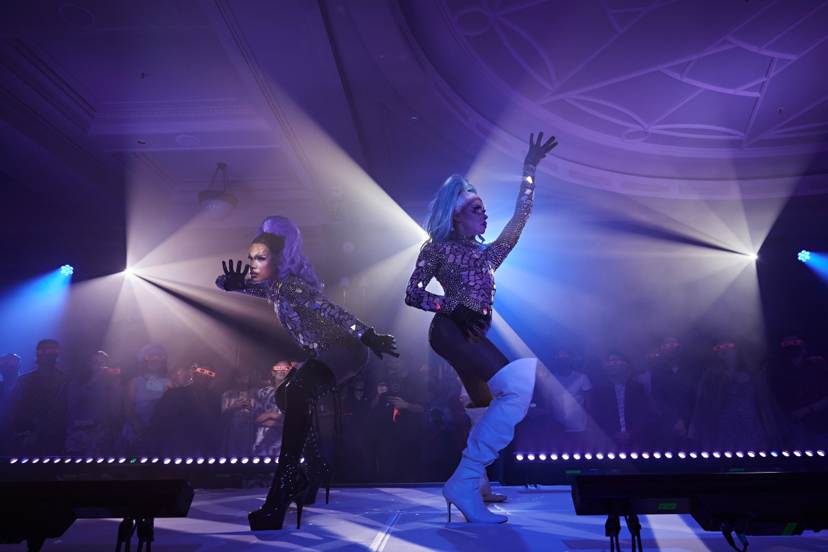RISING: two drag queens in thigh boots, mirrored tops and elaborate plastic look costumes pose on a catwalk among laser purple light beams. There are audience behind them looking up at the catwalk.