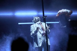 A concert setting, The lead singer, all in white, but with a pink tie, is standing in front of a microphone with their head thrust back.