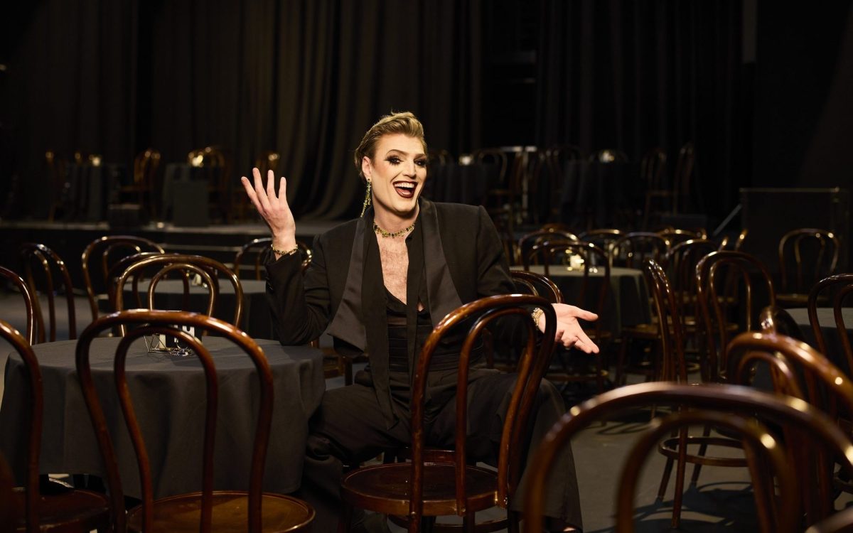 Adelaide Cabaret Festival 2024 Icon Award winner, Reuben Kaye in Space Theatre, Adelaide Festival Centre. Photo: Claudio Raschella. Kaye is in a black suit set with a low neckline, wearing heavy makeup and his hair sleeked back. He is sitting down with a big smile and gesturing with his hands.