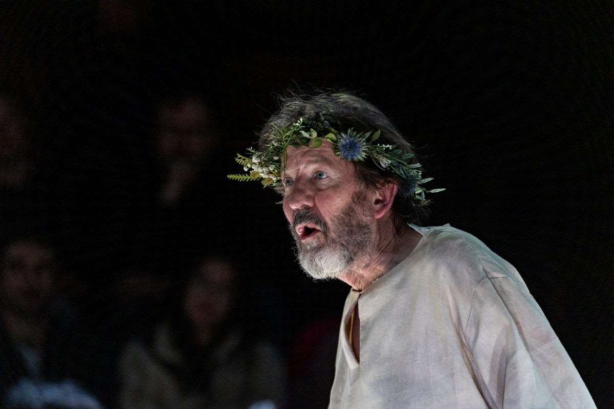 A man with a salt and pepper beard is standing to one side and looking into the distance. He is wearing a white tunic and a crown made of flowers and leaves.